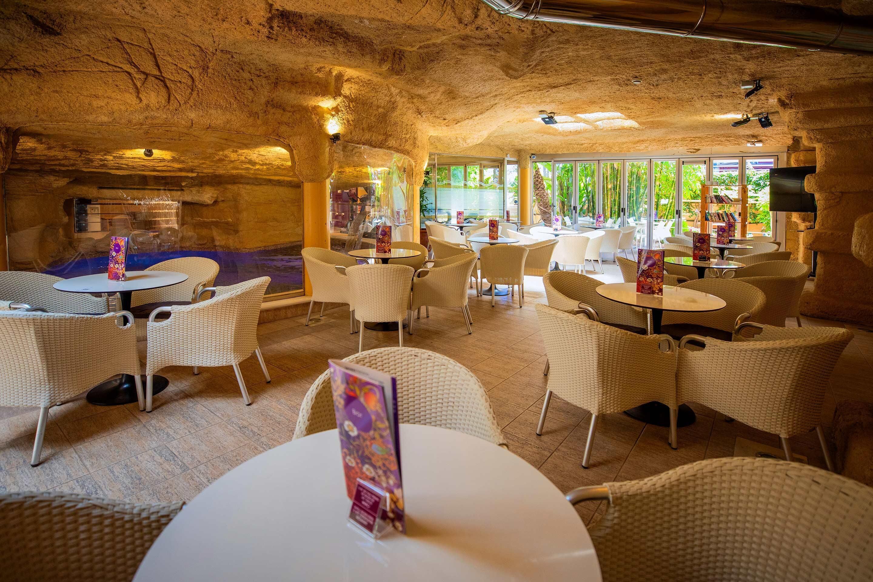 Un espace de restauration intérieur avec des tables rondes et des chaises en osier, doté d’un plafond rappelant une grotte et de grandes fenêtres offrant une vue sur la verdure environnante.