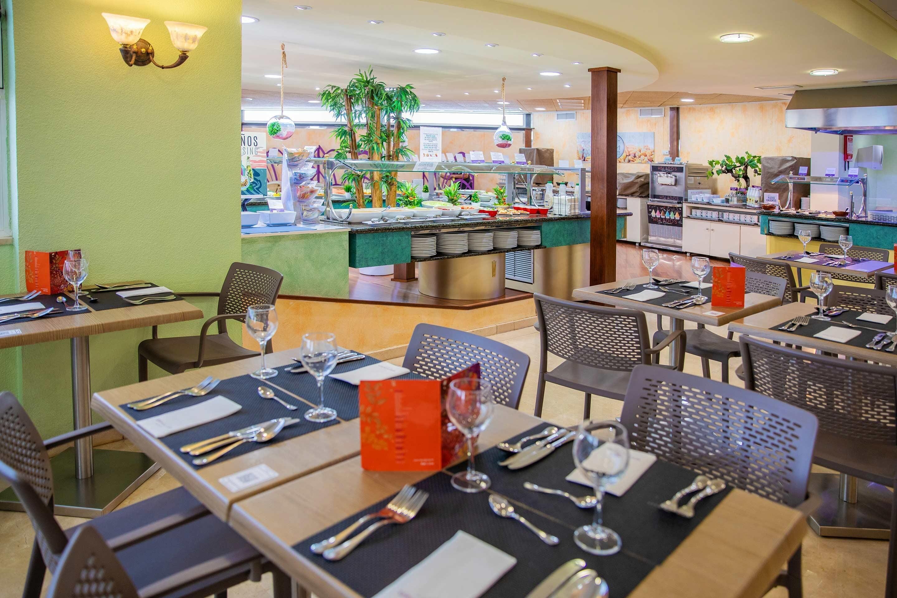 A restaurant with multiple tables set for dining, a buffet area in the background, and a kitchen visible through a glass partition.