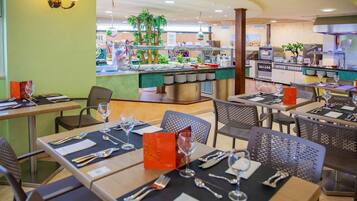 A restaurant with multiple tables set for dining, a buffet area in the background, and a kitchen visible through a glass partition.