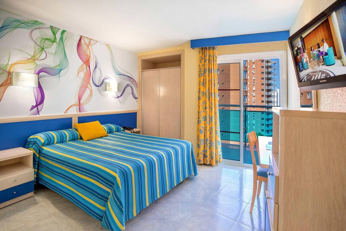 A hotel room with a bed, a desk, a chair, a TV, and a balcony with a view of the city.