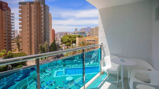 A balcony with a glass railing overlooking a swimming pool area with lounge chairs and a table. - Hotel Servigroup Rialto (Benidorm)