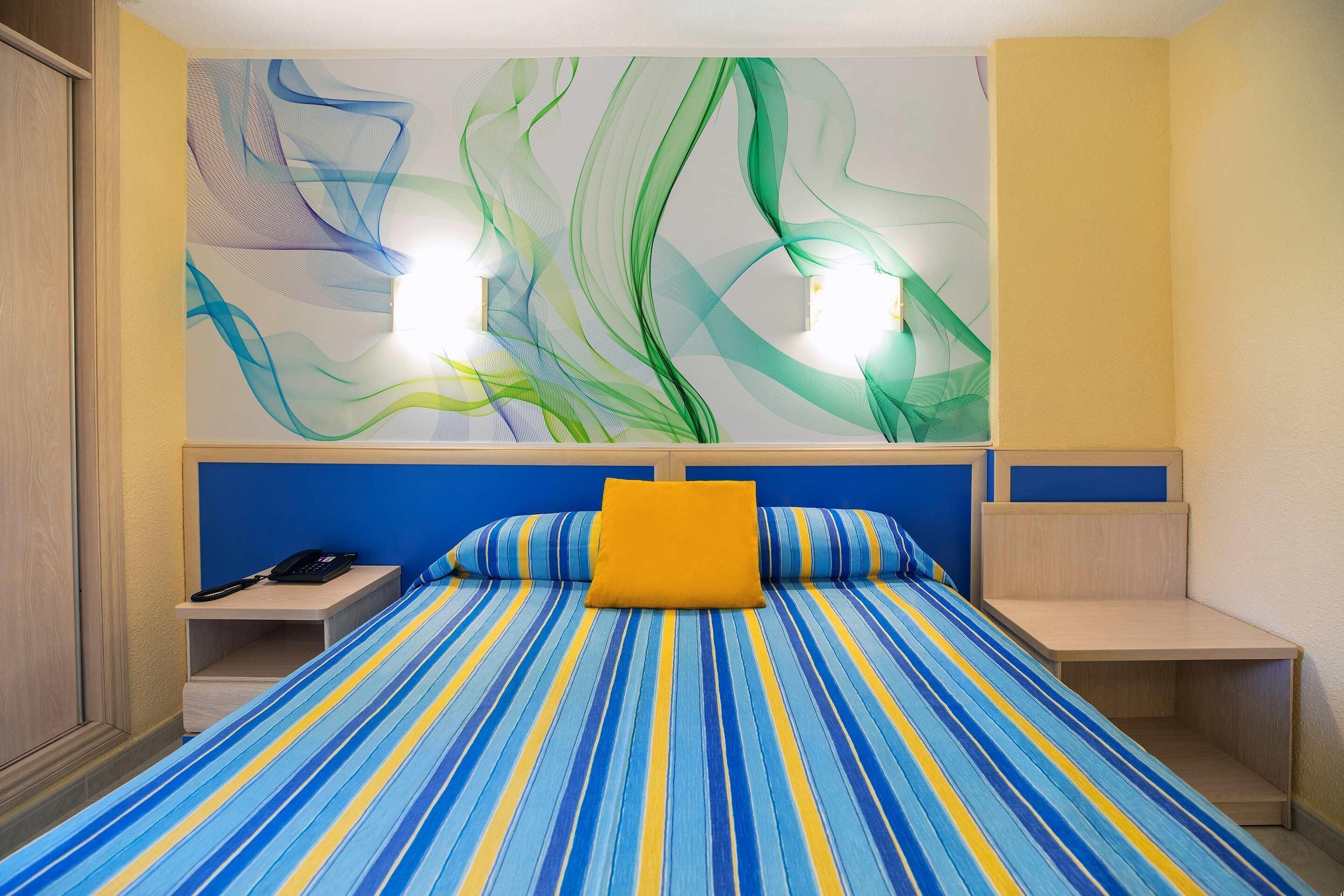 Quarto de hotel com cama coberta por um edredom listrado em azul e amarelo, um travesseiro laranja e cabeceira com painel azul.