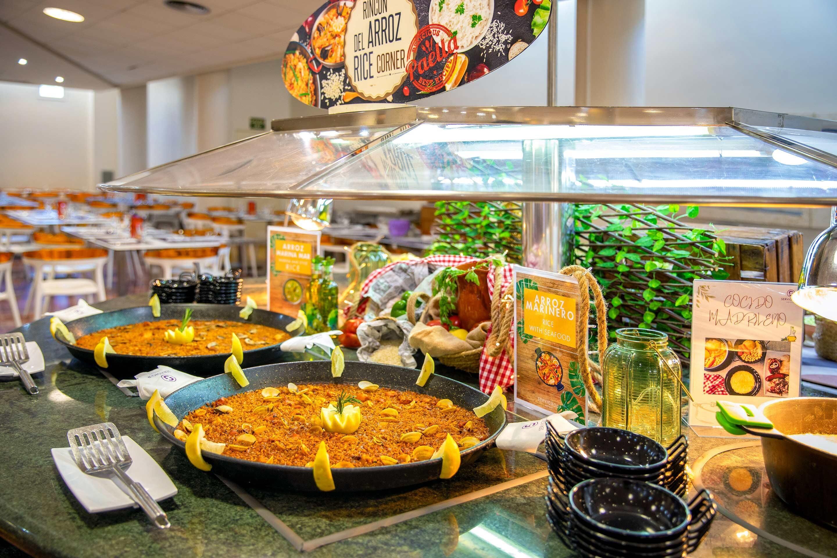 Un bufé con diversos platos, incluyendo arroz y mariscos, y un cartel que dice 'Rincón del Arroz Rice Corner'.