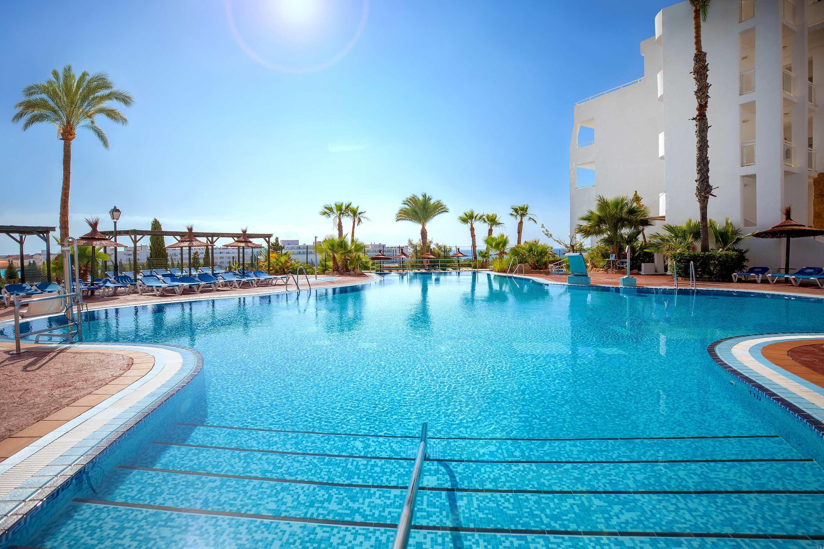 Zona de piscina con sillas de descanso y palmeras, rodeada por un edificio con varios balcones.