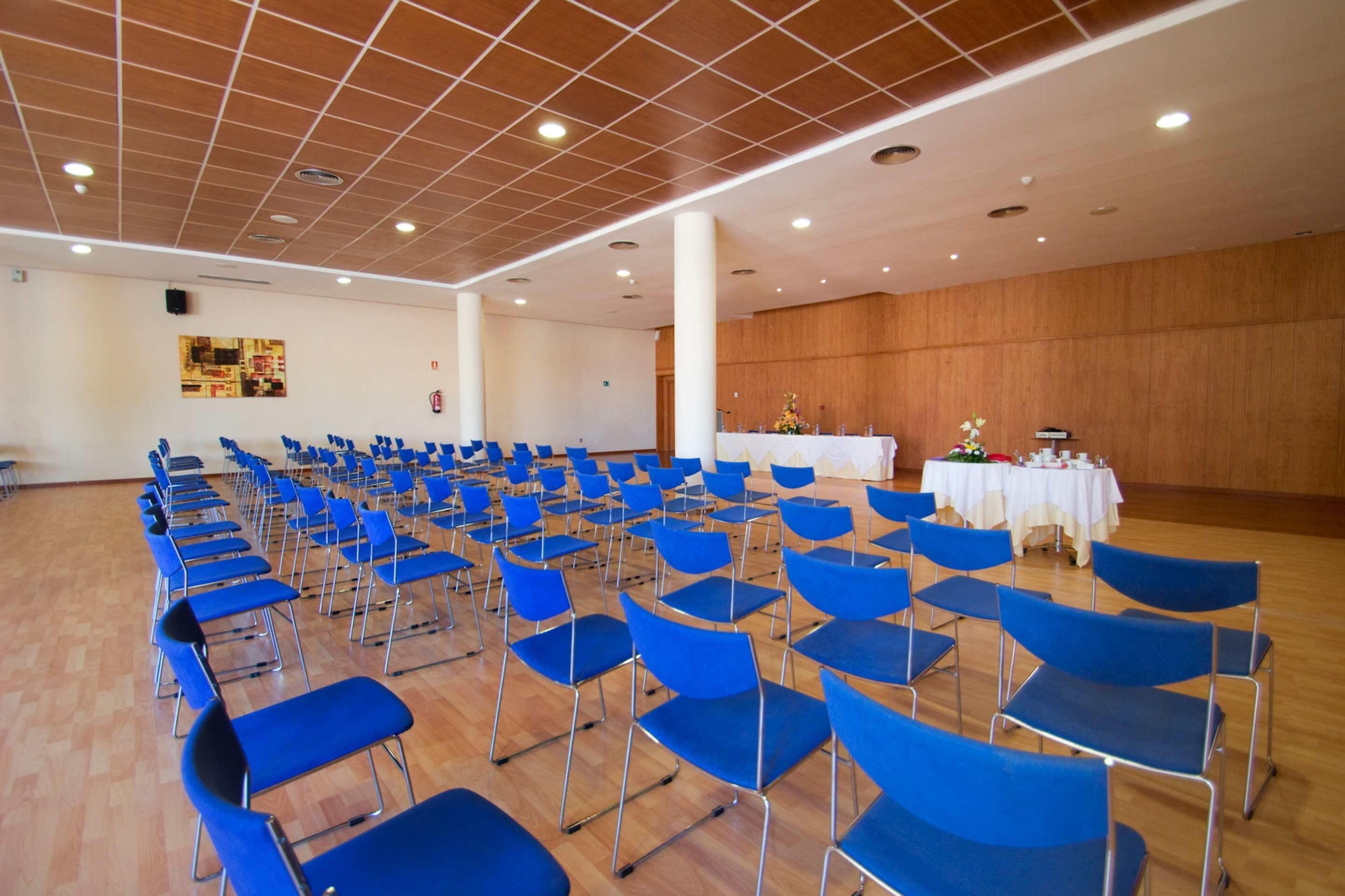 Un amplio salón con filas de sillas y mesas azules, suelo de madera y un panel de pared de madera.