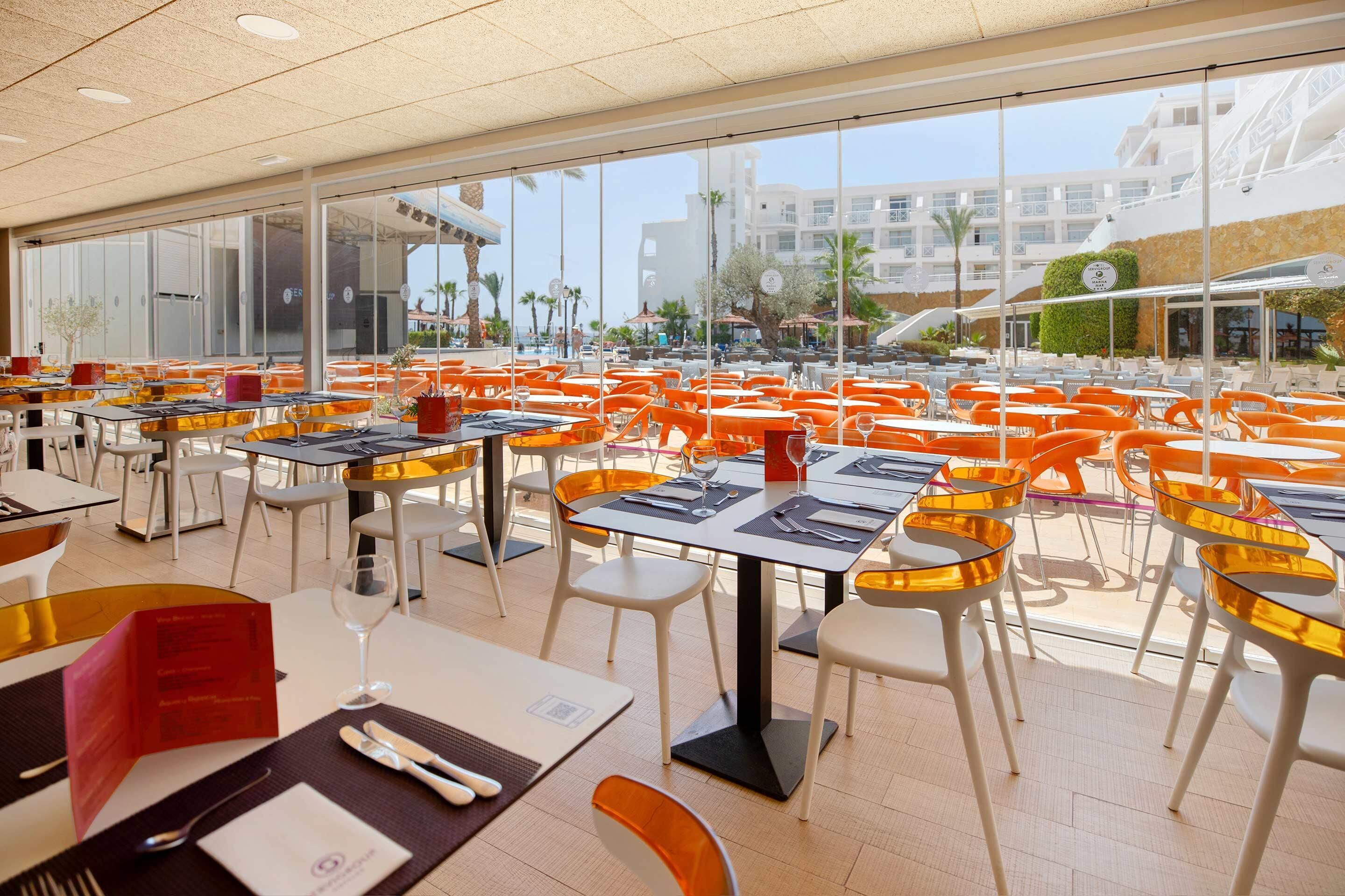 A spacious outdoor dining area with numerous tables and chairs, large windows offering views of the surrounding buildings and palm trees.