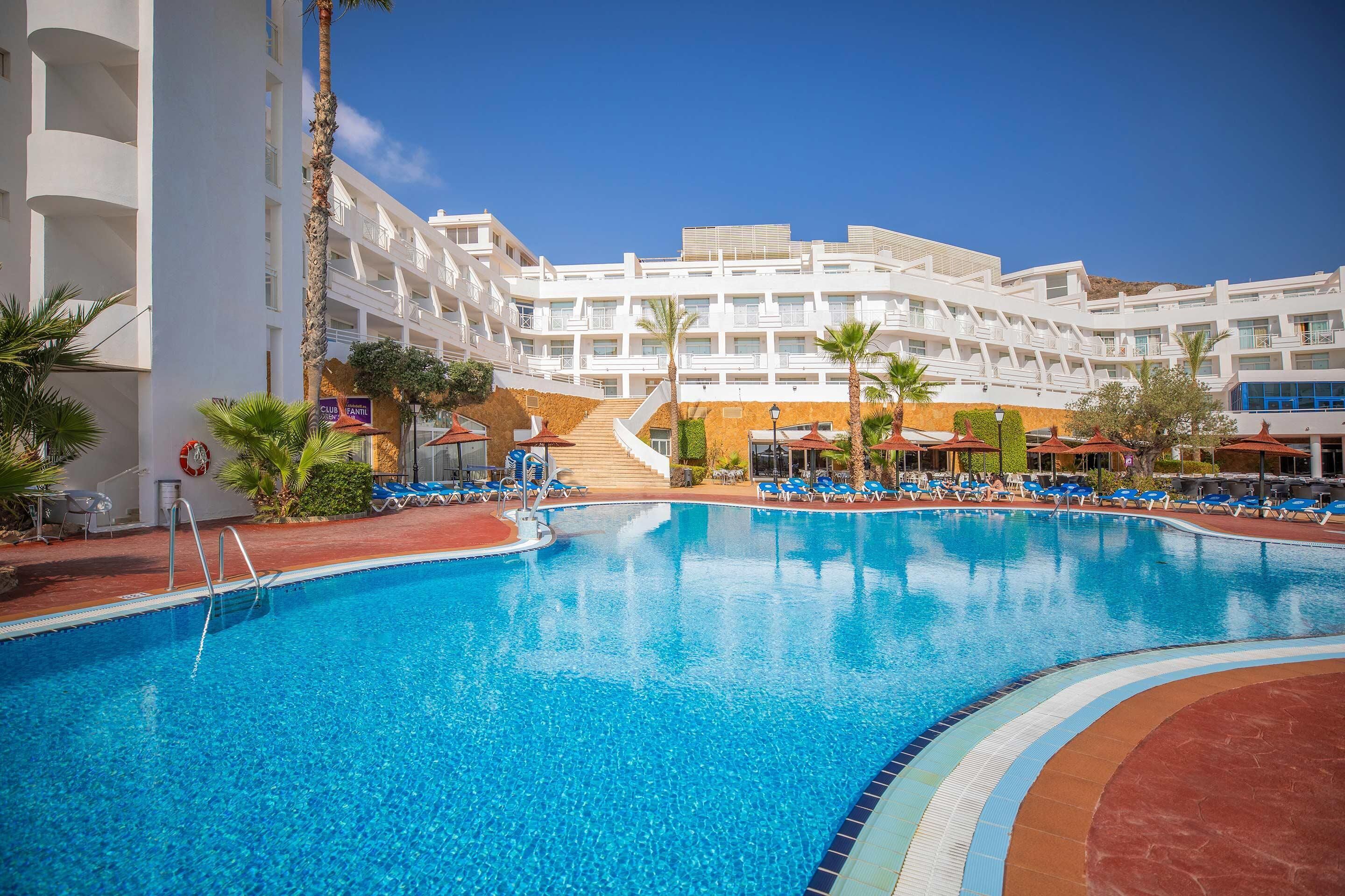 A hotel pool area with a clear blue pool, lounge chairs, and a staircase leading to the upper floors.