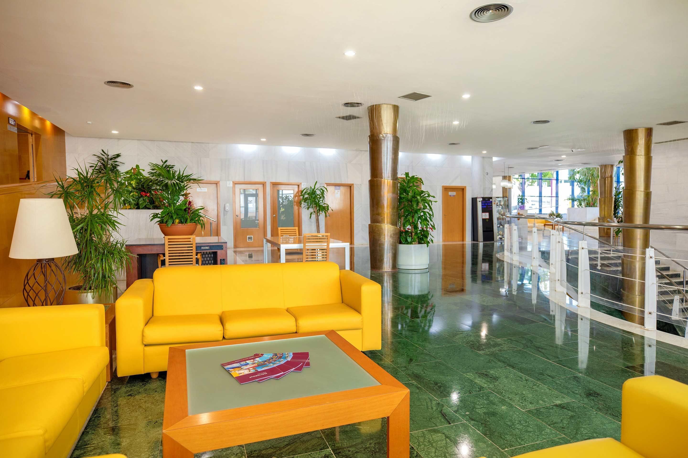 A lobby with a yellow sofa, a coffee table, and potted plants.