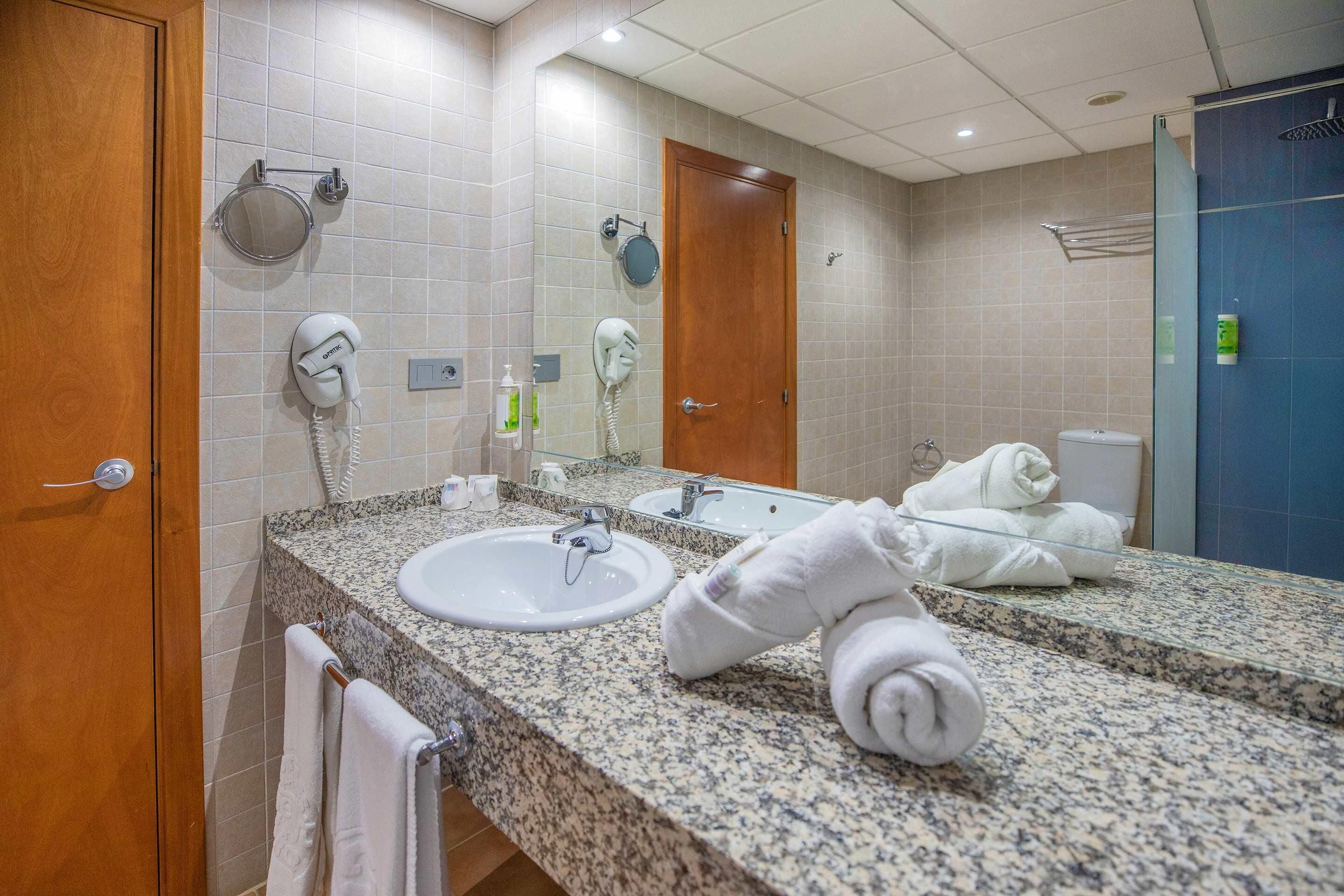 A hotel bathroom with a granite countertop, two sinks, a mirror, a hairdryer, and a towel rack.