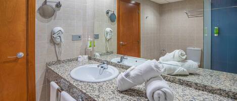 A hotel bathroom with a granite countertop, two sinks, a mirror, a hairdryer, and a towel rack.