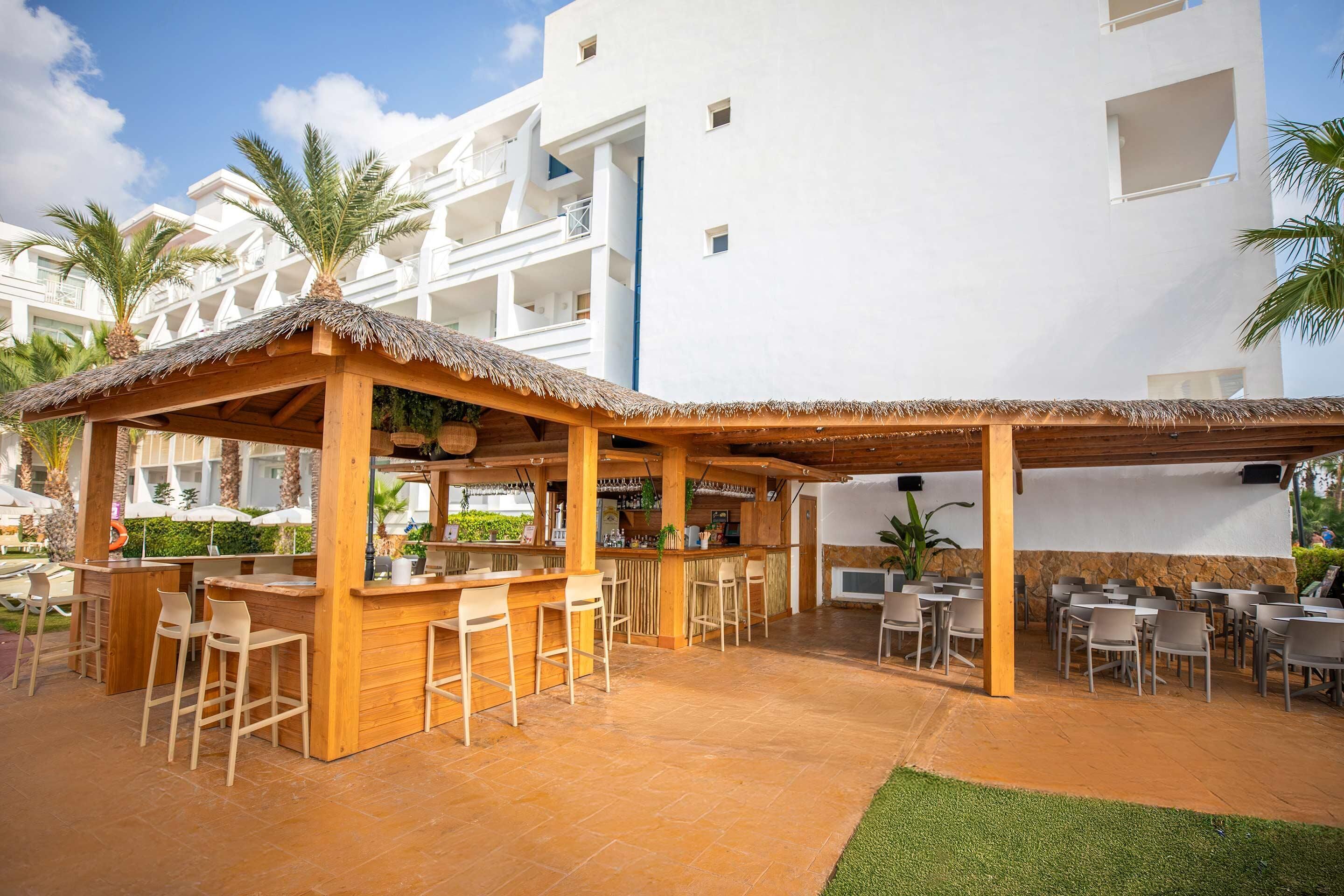 Un área de bar al aire libre, cubierta con taburetes de madera y techo de paja, rodeada de palmeras y un edificio blanco al fondo.