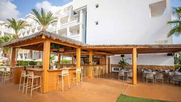 Un área de bar al aire libre, cubierta con taburetes de madera y techo de paja, rodeada de palmeras y un edificio blanco al fondo.