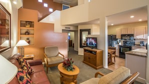 A modern living room with a sofa, armchair, and a small table with a vase. There is a kitchen area with cabinets and a refrigerator. - Villas of Cave Creek (Cave Creek)