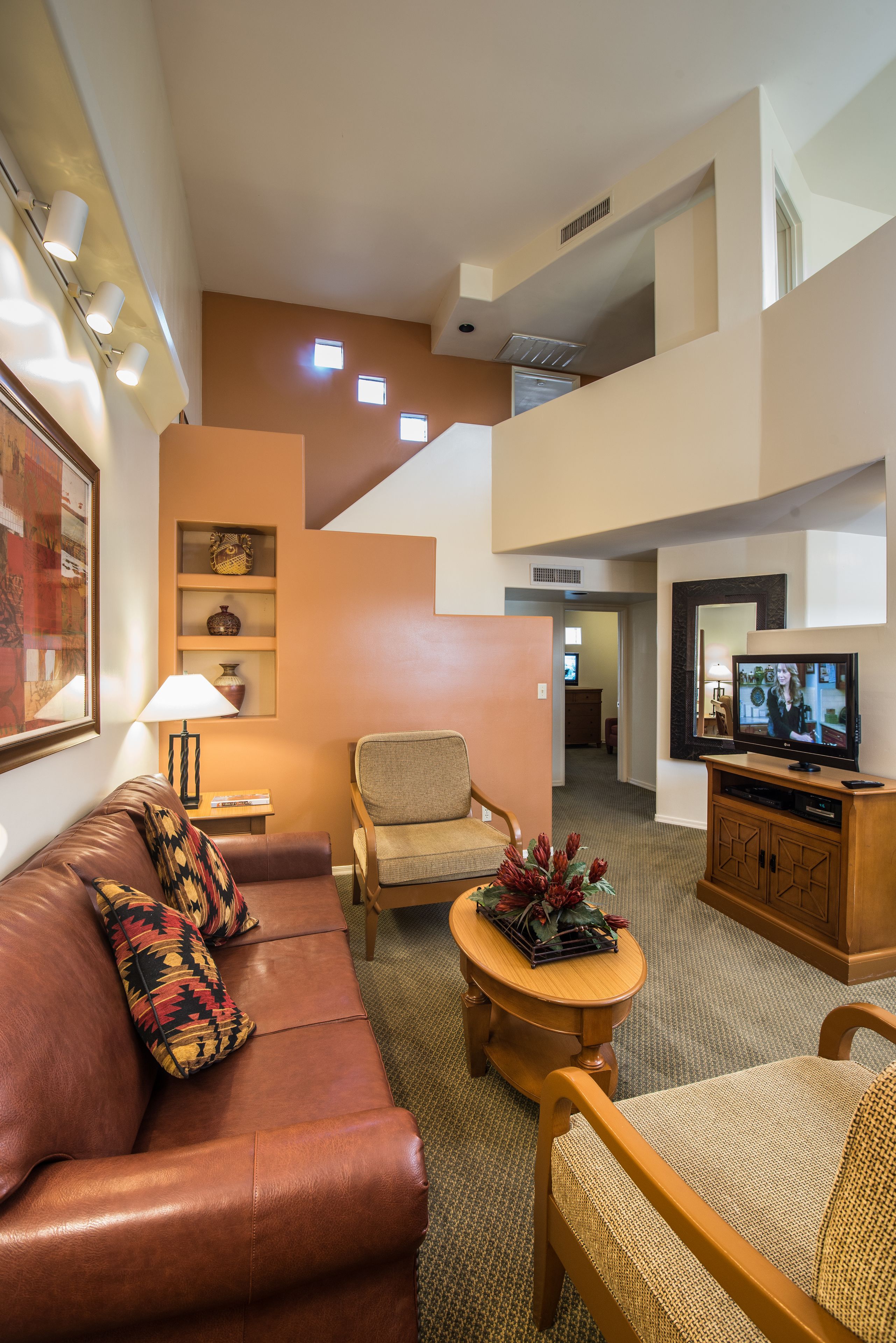 A modern living room with a brown leather sofa, a wooden coffee table, and a television on a wooden stand. There is a staircase leading to an upper level.
