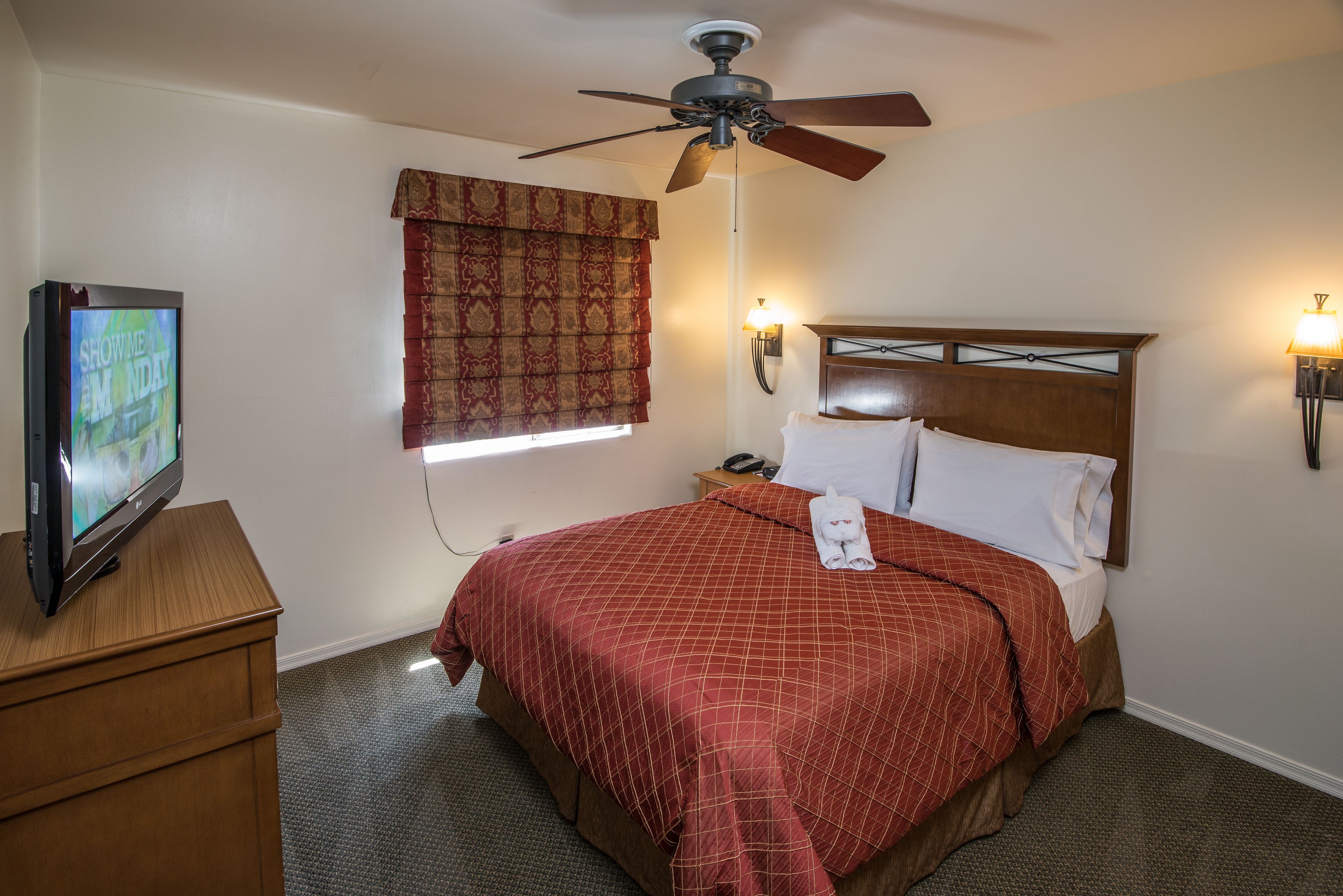 Quarto de hotel com cama, televisão, ventoinha no teto, mesa de cabeceira e janela com cortinas.