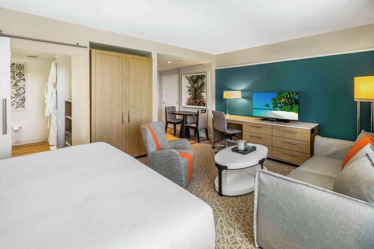 A hotel room with a large bed, a sofa, a desk with a computer, a TV, and a bathroom with a shower and a towel rack.
