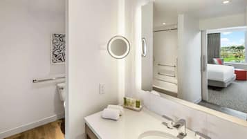Un baño moderno con un espejo grande, un lavabo y un inodoro. Hay un perchero con toallas y una pequeña bandeja con artículos de aseo. El baño tiene vista a una cama y una ventana.
