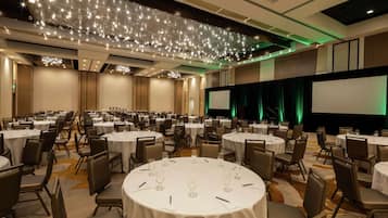 Un amplio salón de banquetes con mesas redondas, sillas, un escenario con una pantalla y un techo decorado con luces que parecen estrellas.