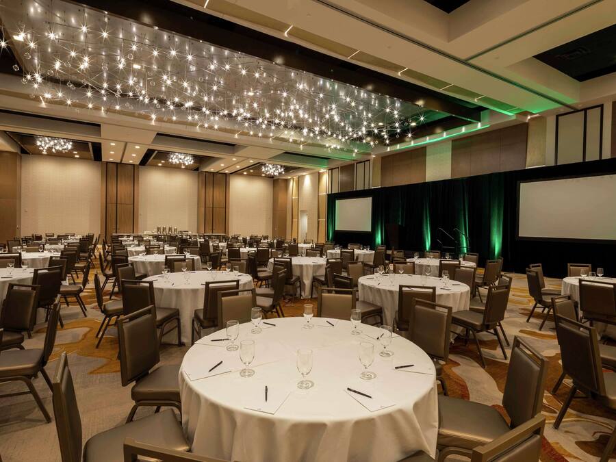 Un amplio salón de banquetes con mesas redondas, sillas, un escenario con una pantalla y un techo decorado con luces que parecen estrellas.