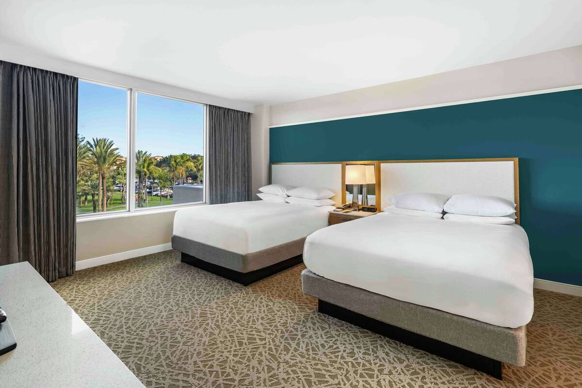 A hotel room with two beds, a large window with a view of palm trees, a desk, and a patterned carpet.