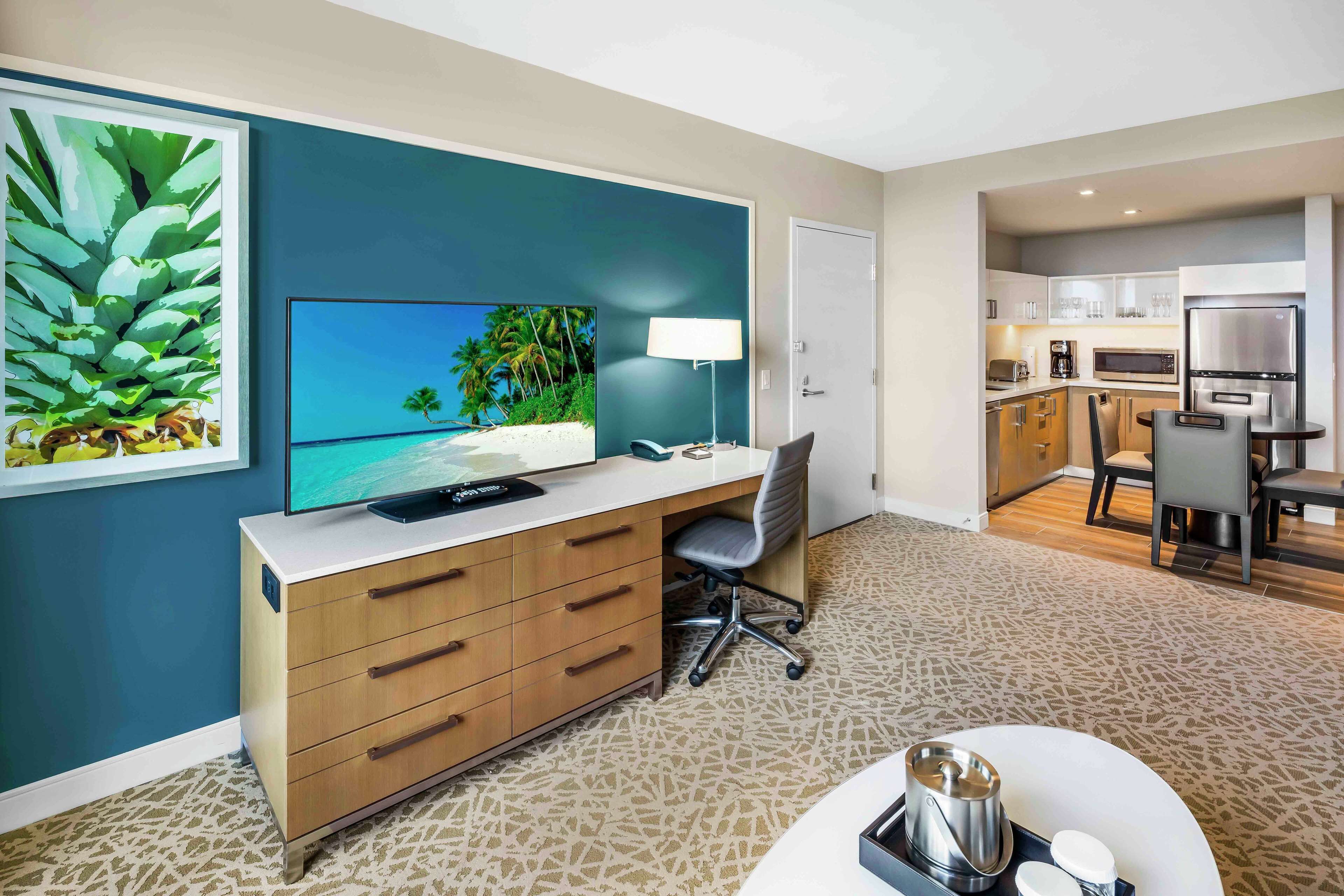 Quarto de hotel moderno com televisão, mesa de trabalho e cozinha compacta.