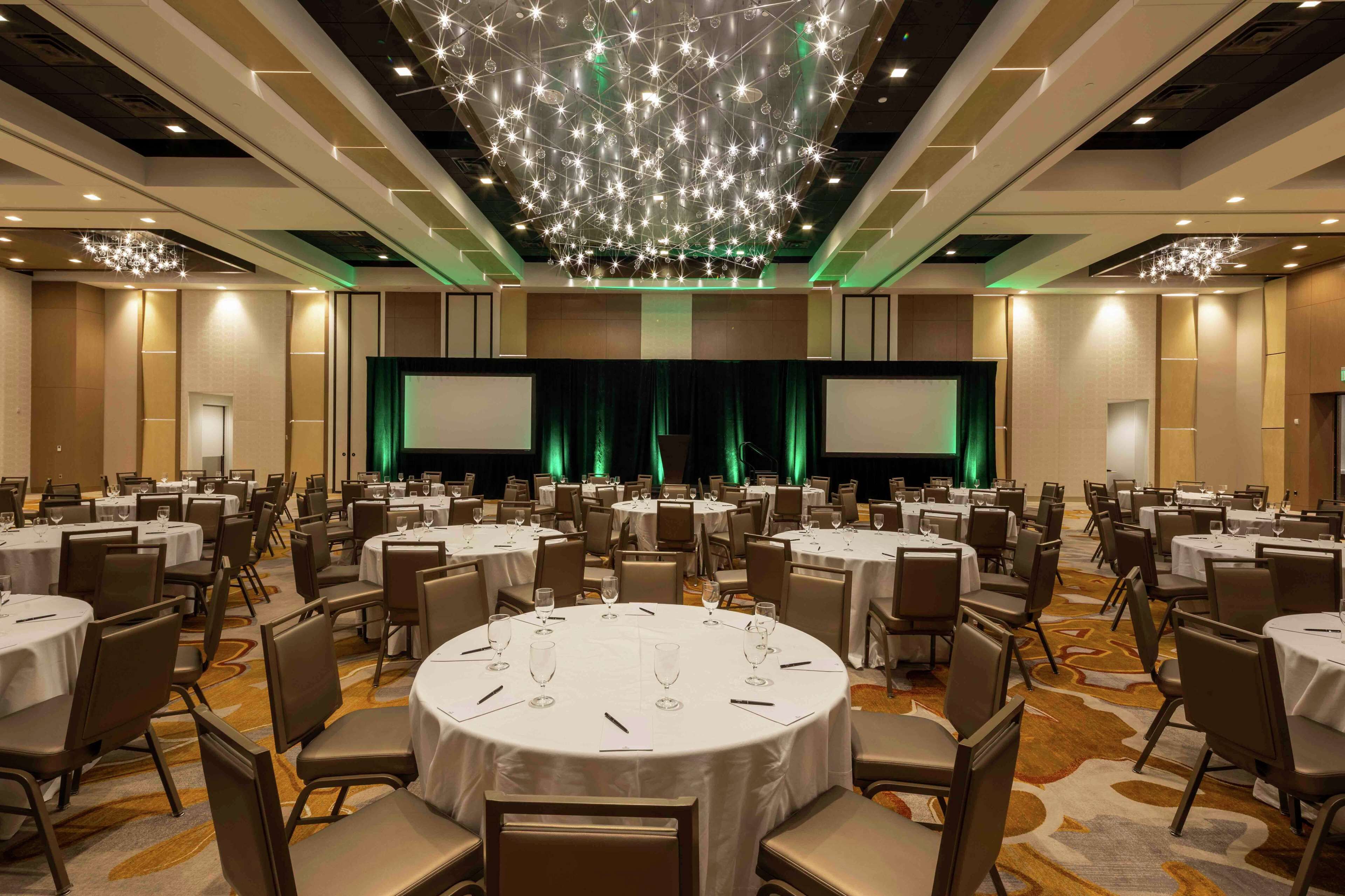 A large conference room with round tables, chairs, and a stage with a green curtain.