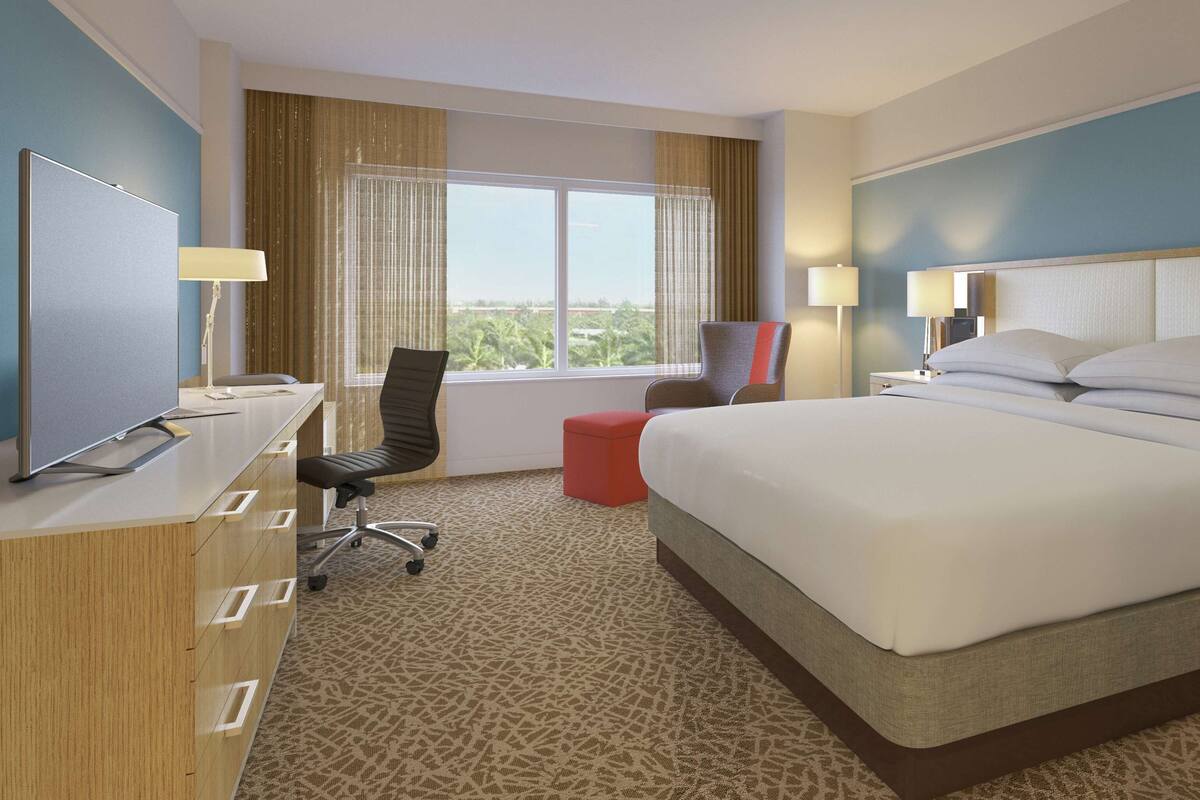 A hotel room with a large bed, a desk with a computer, a chair, and a window with curtains.