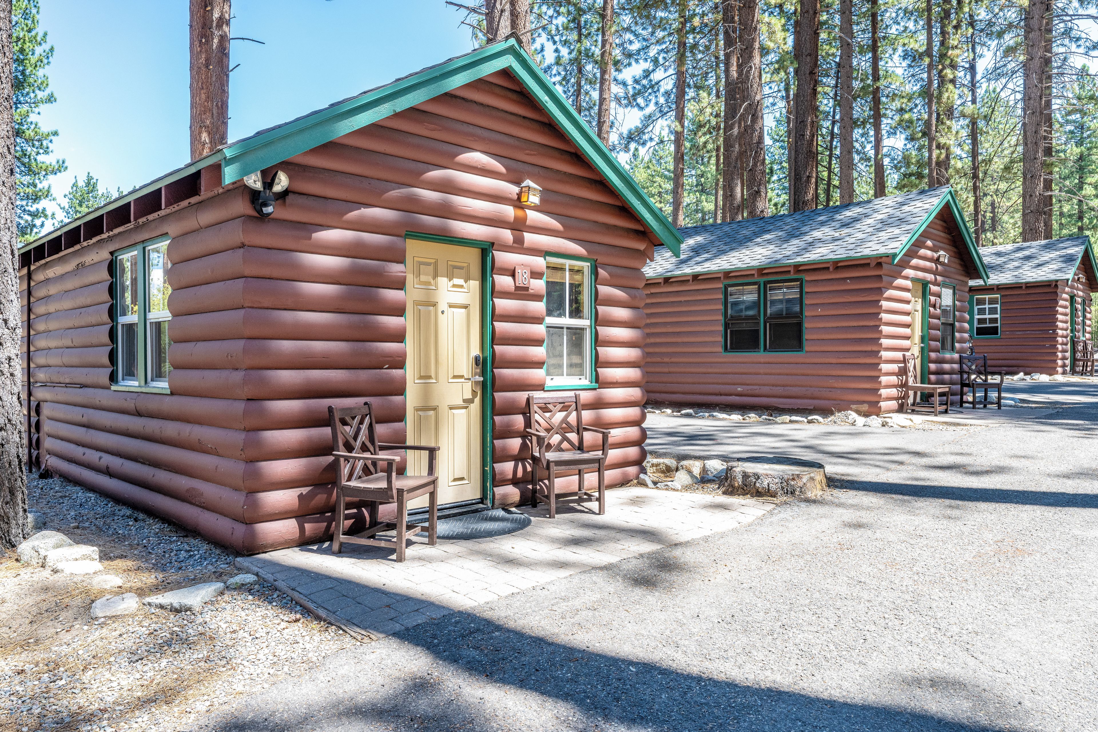A log cabin with a green roof, a front door, and two windows. There are two wooden chairs outside.