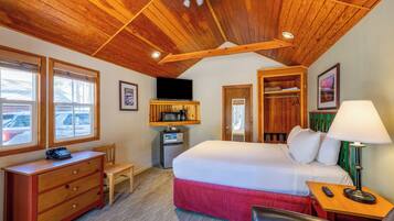A hotel room with a wooden ceiling, a bed with a red bedspread, a desk with a telephone, a chair, a small refrigerator, a microwave, and a television.