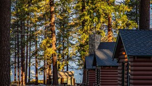 A cabin-style lodge with a stone chimney, wooden benches, and a view of tall trees and a body of water.
