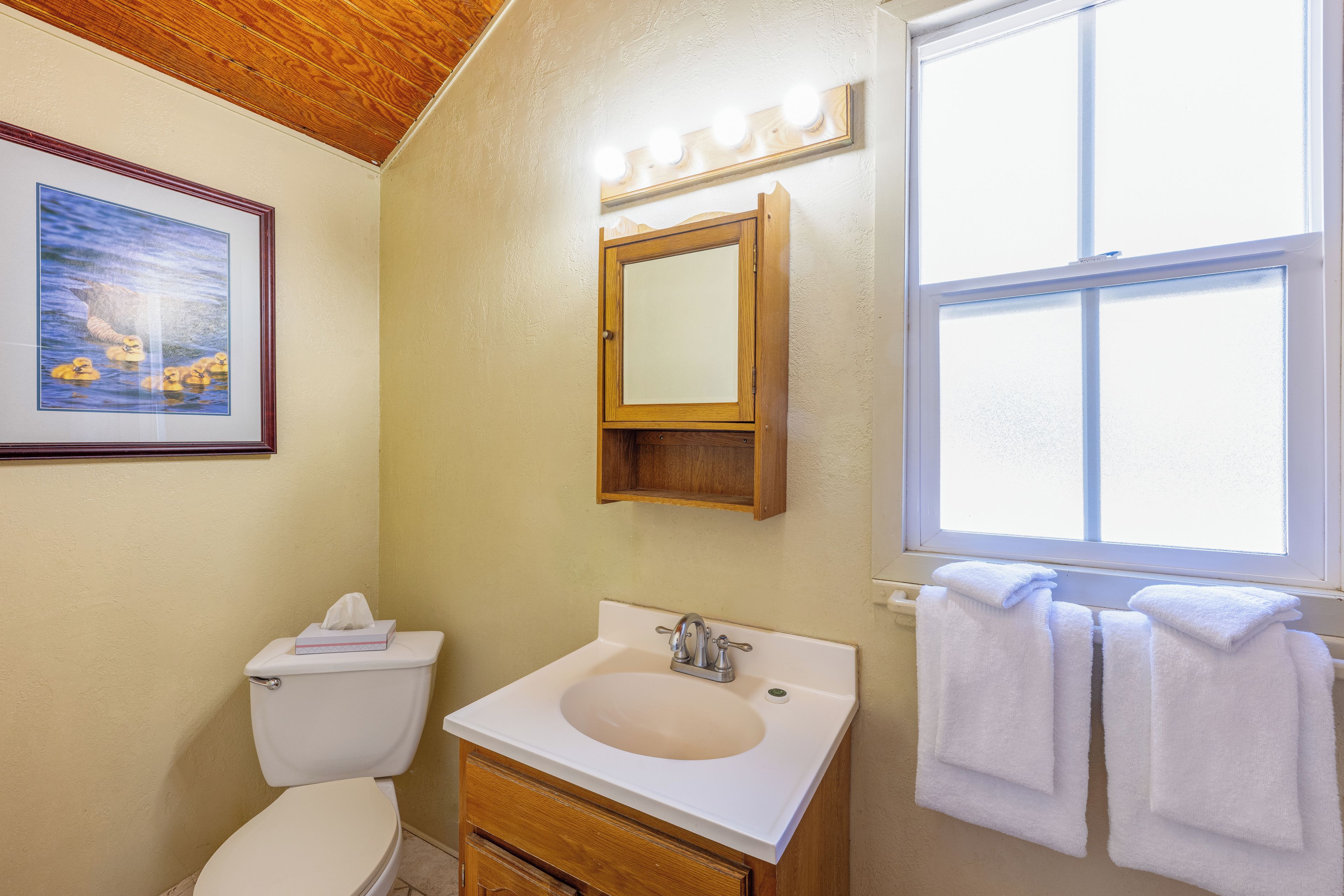 A bathroom with a toilet, sink, mirror, and a window.