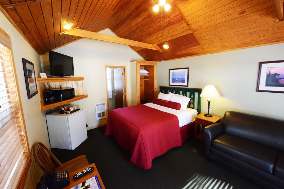 A hotel room with a wooden ceiling, a bed with red bedding, a black leather sofa, a television on a shelf, and a small refrigerator.