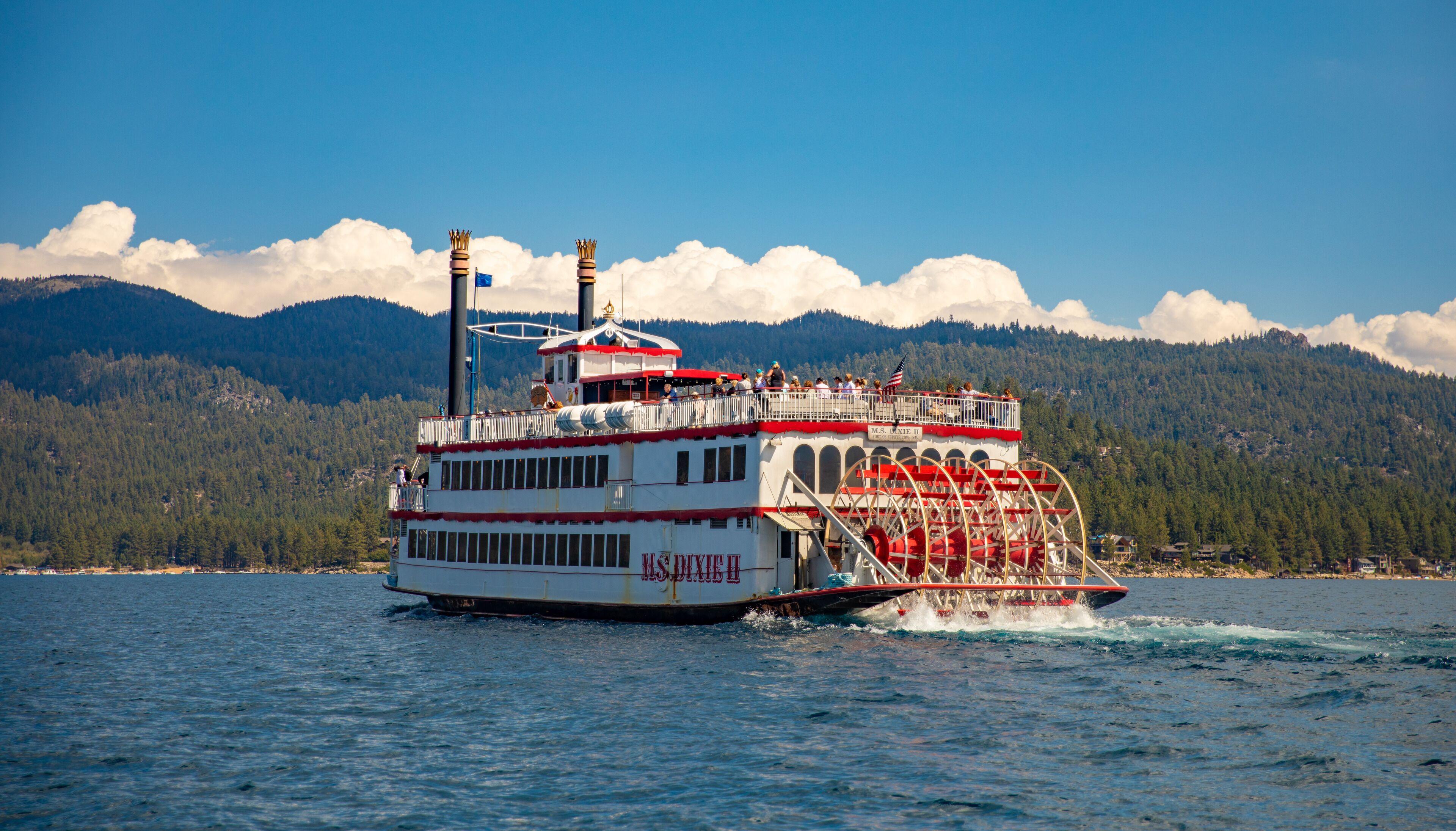 A large paddle steamer with multiple decks and a prominent wheelhouse, navigating on a body of water with forested mountains in the background.