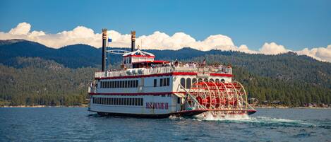 A large paddle steamer with multiple decks and a prominent wheelhouse, navigating on a body of water with forested mountains in the background.