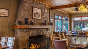 A rustic interior with a stone fireplace, wooden furniture, and a view through large windows.