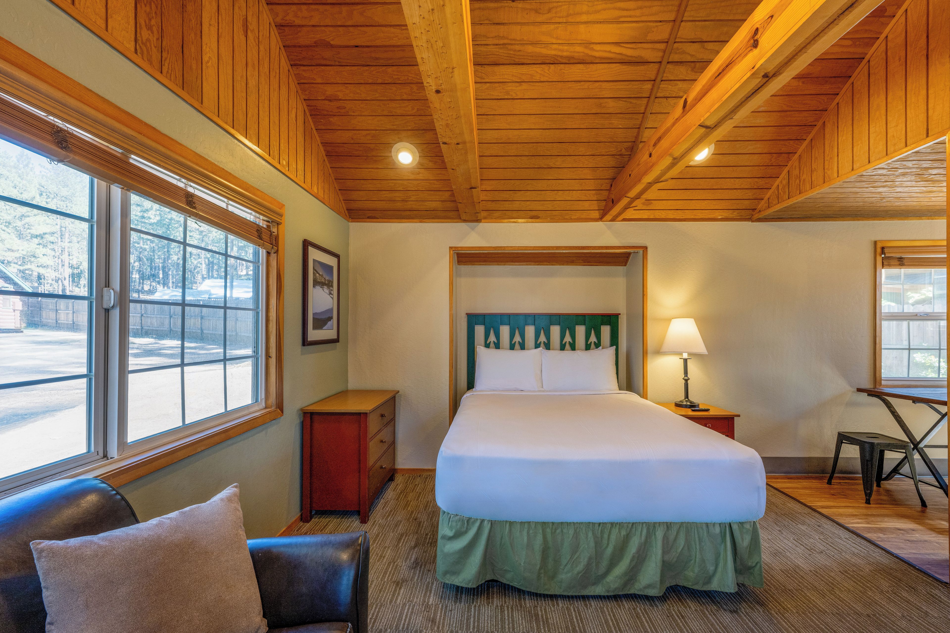 A hotel room with a large bed, a nightstand, a lamp, a chair, and a window with a view of trees.