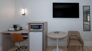 A compact hotel room with a kitchenette, a desk, a chair, a small refrigerator, a microwave, a television, and a round table.