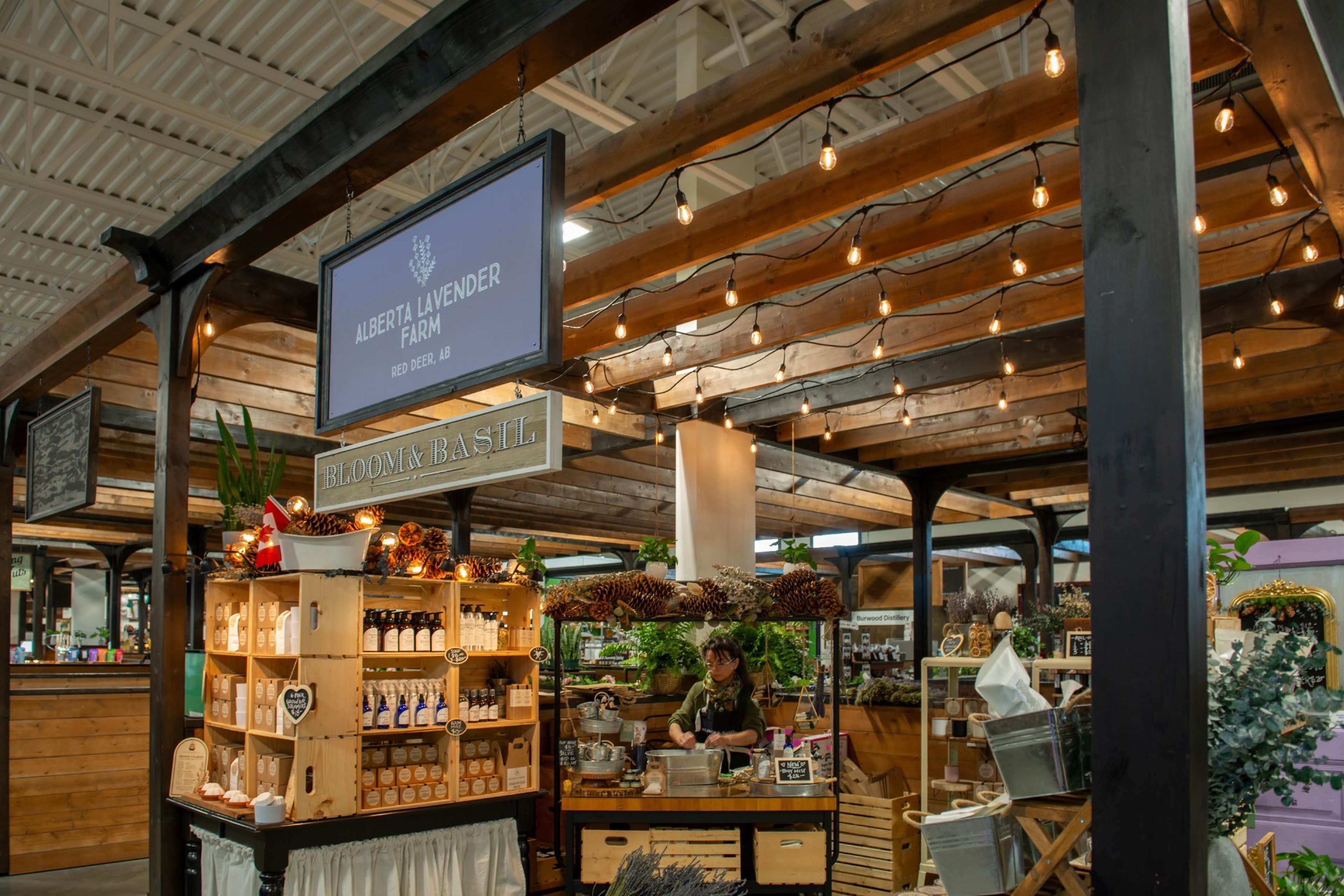 A farm-to-table market stall with wooden shelves stocked with products, a sign indicating Alberta Lavender Farm, and a rustic interior with exposed beams and string lights.