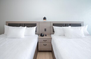Habitación de hotel con dos camas, cada una con ropa blanca y almohadas, una mesita de noche con teléfono y lámpara, e interruptor de luz en la pared.