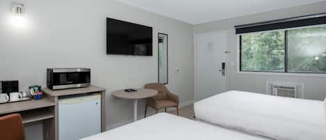 A hotel room with a bed, a desk with a microwave, a chair, and a window with a view of trees.