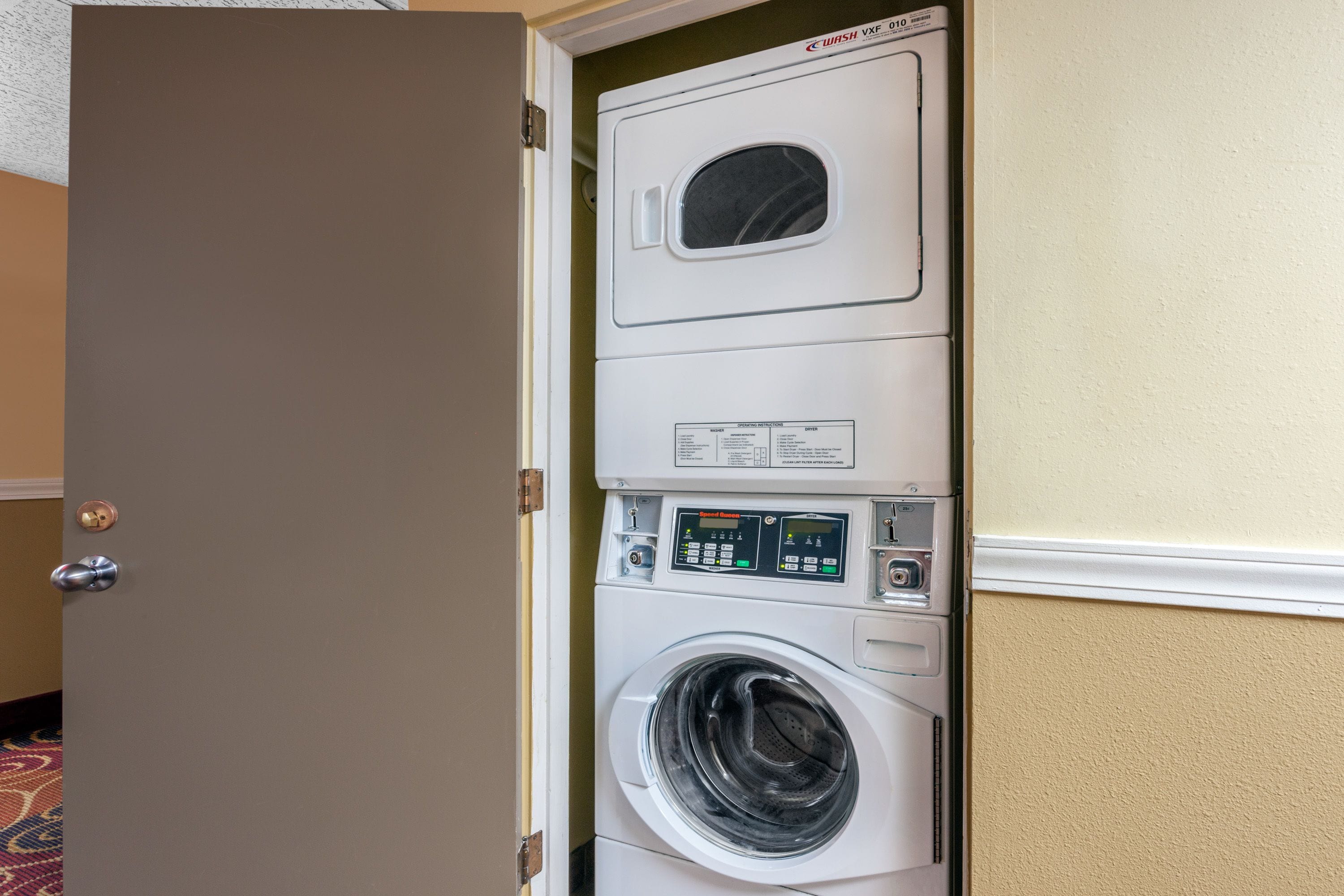 Una lavadora y secadora apiladas con un panel de control, ubicadas en una habitación con paredes color beige y alfombra estampada.