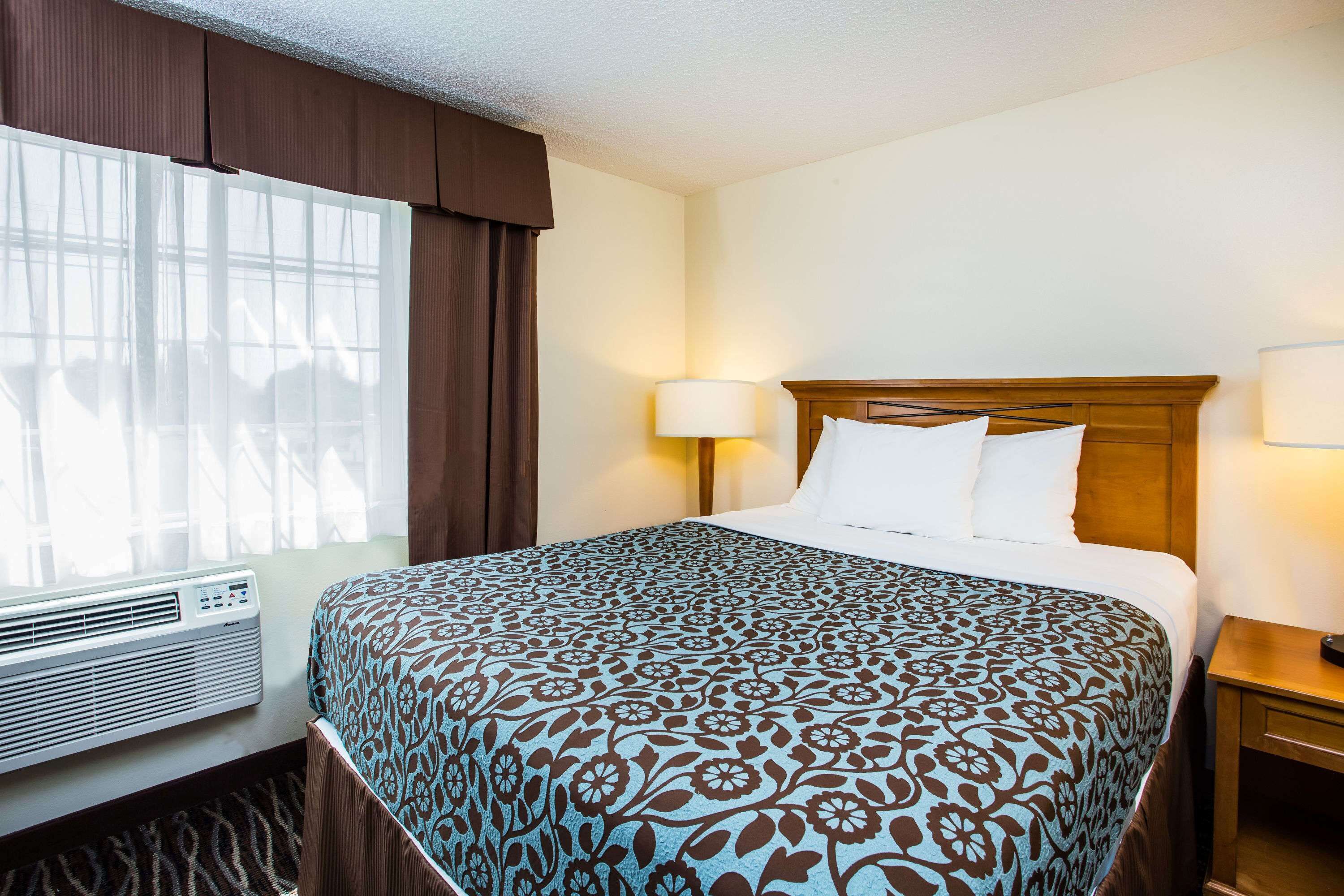 A hotel room with a large bed, a window with curtains, a wall-mounted air conditioner, and a bedside lamp.