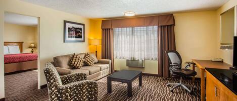 A hotel room with a sofa, armchair, and desk. There is a bed with a red headboard in the background.