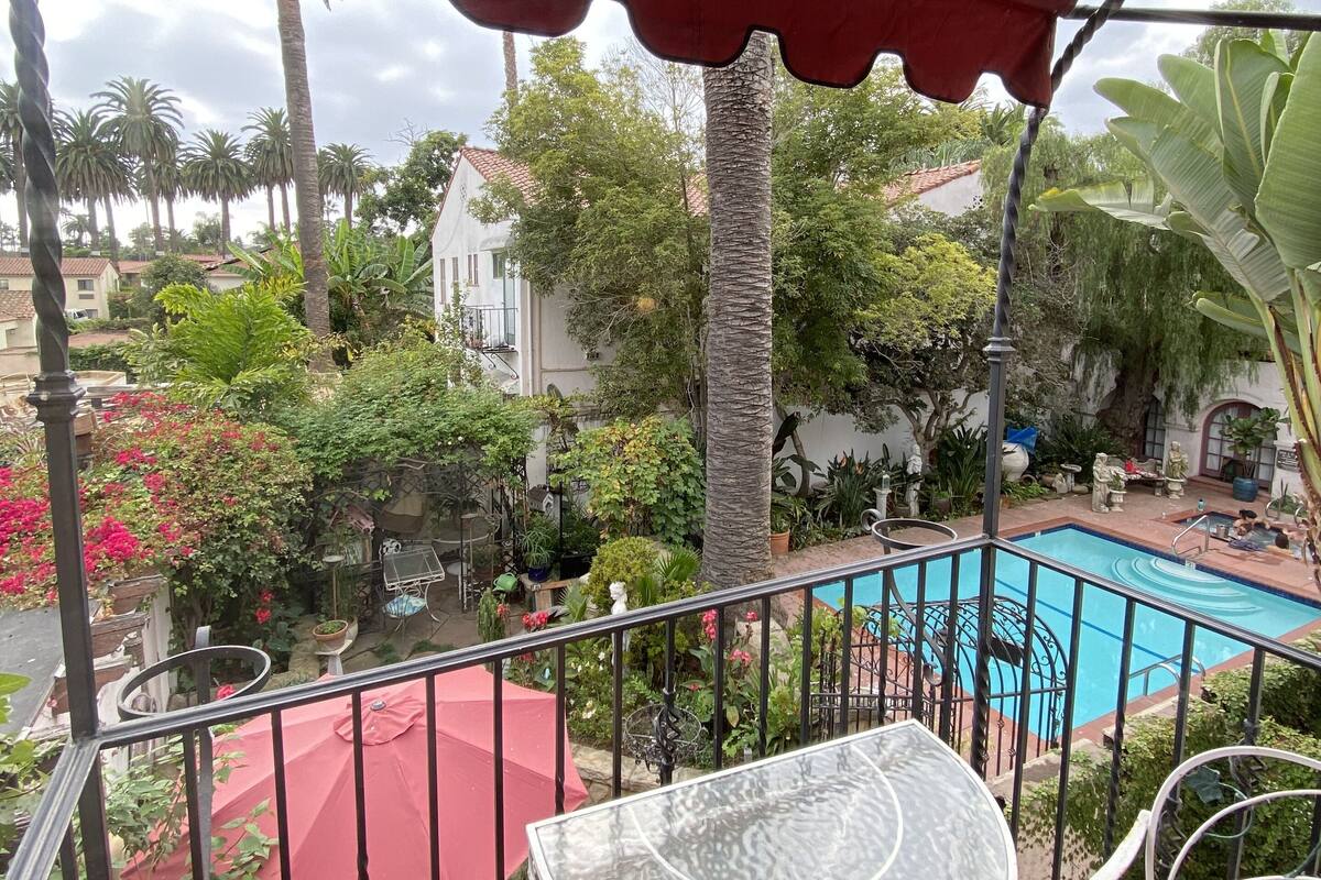 A balcony with a glass table and red cushions, overlooking a swimming pool and lush greenery.