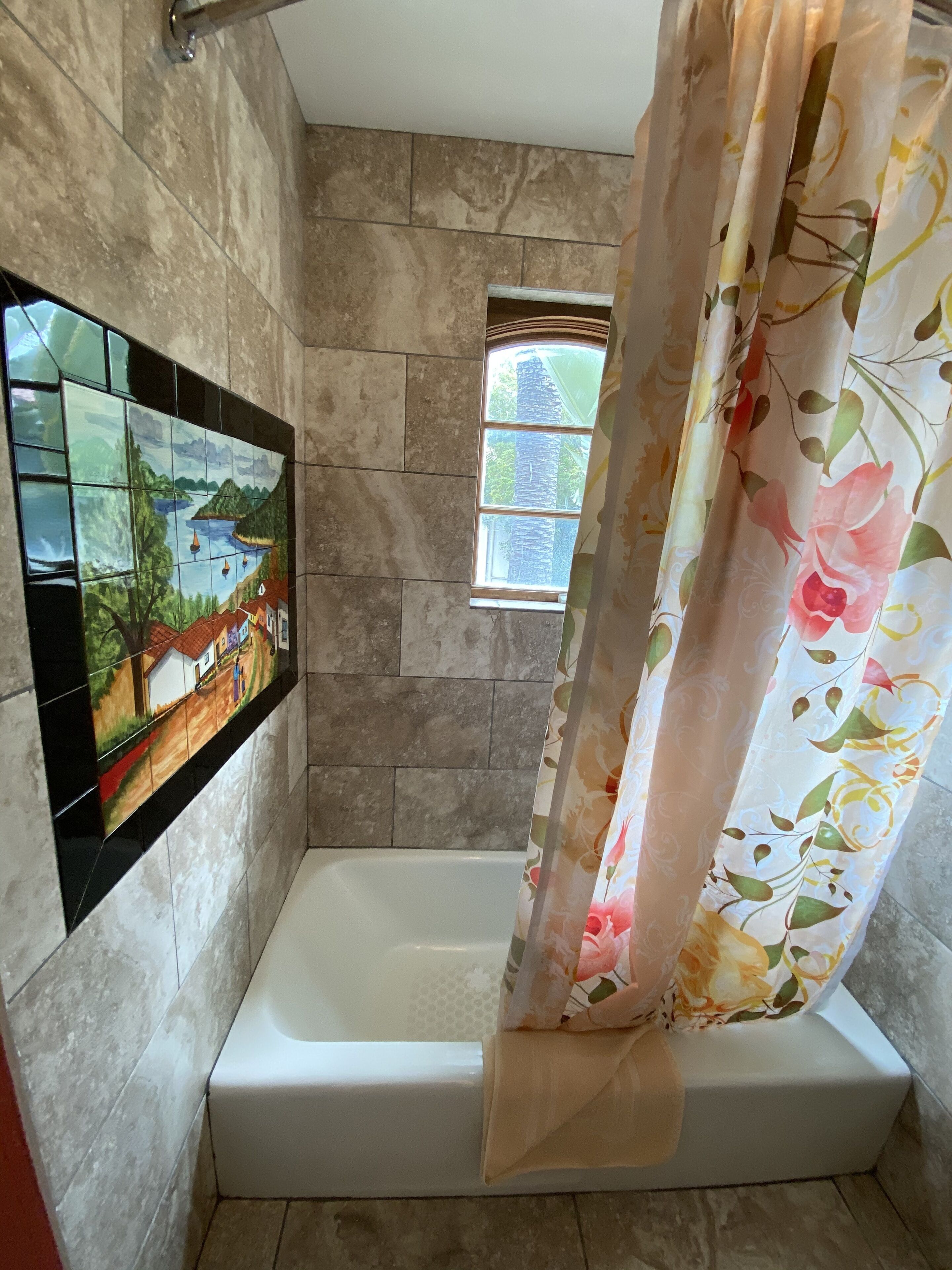 A bathroom with a shower curtain, a small window, and a tiled wall with a mural.