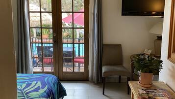 Habitación de hotel con cama, una silla, una mesa con planta, televisor y balcón con piscina y sombrillas.