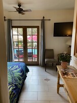 A hotel room with a bed, a chair, a table with a plant, a TV, and a balcony with a pool and umbrellas.