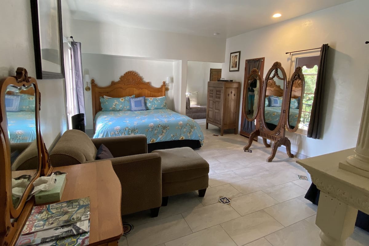 A bedroom with a large bed, a wooden wardrobe, a chair, a small table, and a mirror.
