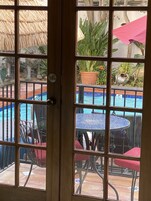 Vista através de uma porta de vidro para uma área de piscina com piso de azulejos, uma mesa redonda com cadeiras e plantas em vasos.