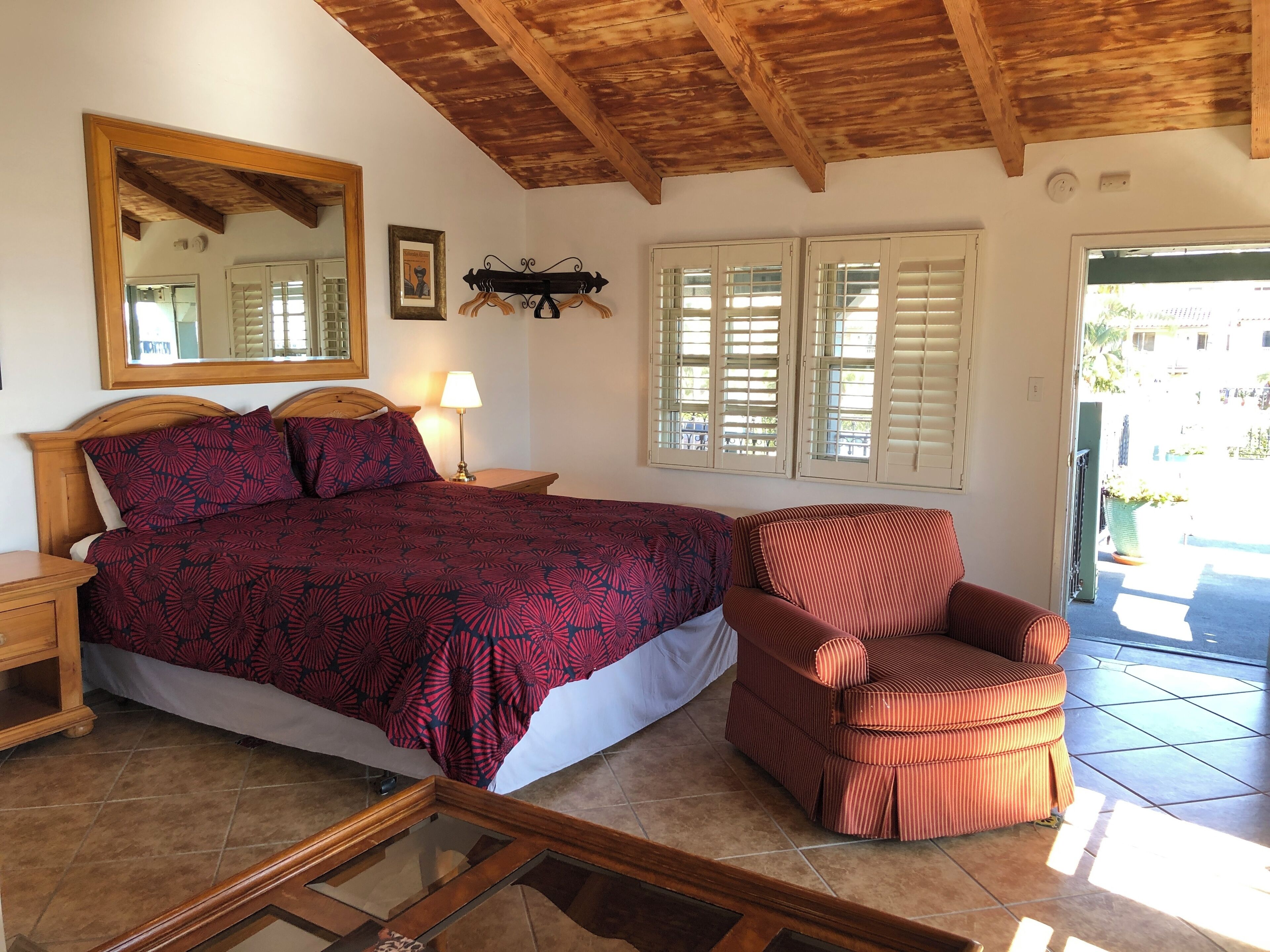 A bedroom with a bed, a chair, a nightstand, a lamp, a mirror, and a window with shutters.